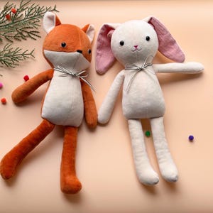May include: Two plush toys, a fox and a bunny, are sitting on a light peach background. The fox is orange and white with black eyes and a black and white ribbon around its neck. The bunny is white with pink ears and a black and white ribbon around its neck.