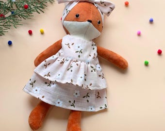 May include: A plush fox doll wearing a pink dress with white flowers and green leaves. The doll has a brown nose and black eyes. The doll is wearing a white headband with a bow.