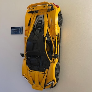May include: A yellow Lego McLaren 720S model car is mounted on a white wall. The car is partially disassembled, showing the inner workings of the model. The car is labeled "McLaren 720S" on a blue plaque.