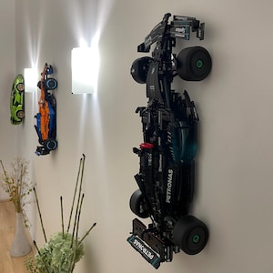 Peut inclure: Un modèle de voiture de course de Formule 1 Lego noir et vert avec la marque Petronas est monté sur un mur blanc. La voiture est à l'envers, les roues face au spectateur. La voiture est une réplique de la voiture de l'équipe Mercedes-AMG Petronas de Formule 1.