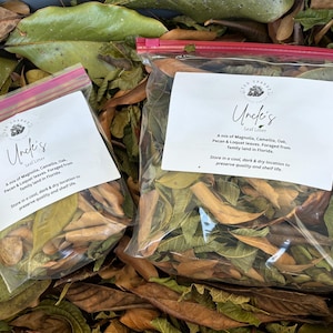 May include: Two clear plastic bags filled with dried leaves, labeled "Uncle's Leaf Litter." The bags are sealed with pink zippers and rest on a bed of fallen leaves. The labels list the leaf mix and origin.