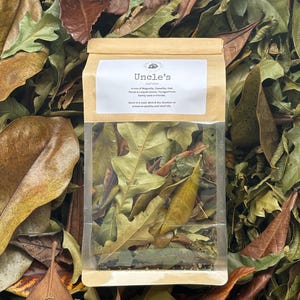 May include: A clear plastic and brown paper pouch filled with a mix of dried Magnolia, Camellia, Oak, Pecan, and Loquat leaves. The label reads Uncle's Leaf Litter, A mix of Magnolia, Camellia, Oak, Pecan & Loquat leaves. Foraged from family land in Florida.
