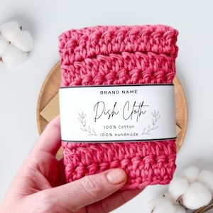 May include: A handmade, hot pink dish cloth made of 100% cotton. The cloth is wrapped with a white label that reads "Dish Cloth" with the brand name and details about the material. The cloth is held in a person's hand.