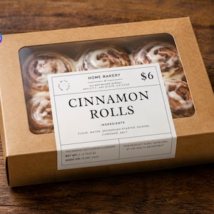 May include: A rectangular brown cardboard box with a clear window containing six cinnamon rolls topped with white icing. A white label on the front reads HOME BAKERY, CINNAMON ROLLS, ingredients, and net weight 7 oz (200 g) with a date of 12/08/2026.