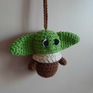May include: A green crocheted Yoda ornament with brown and white details hanging from a brown string.