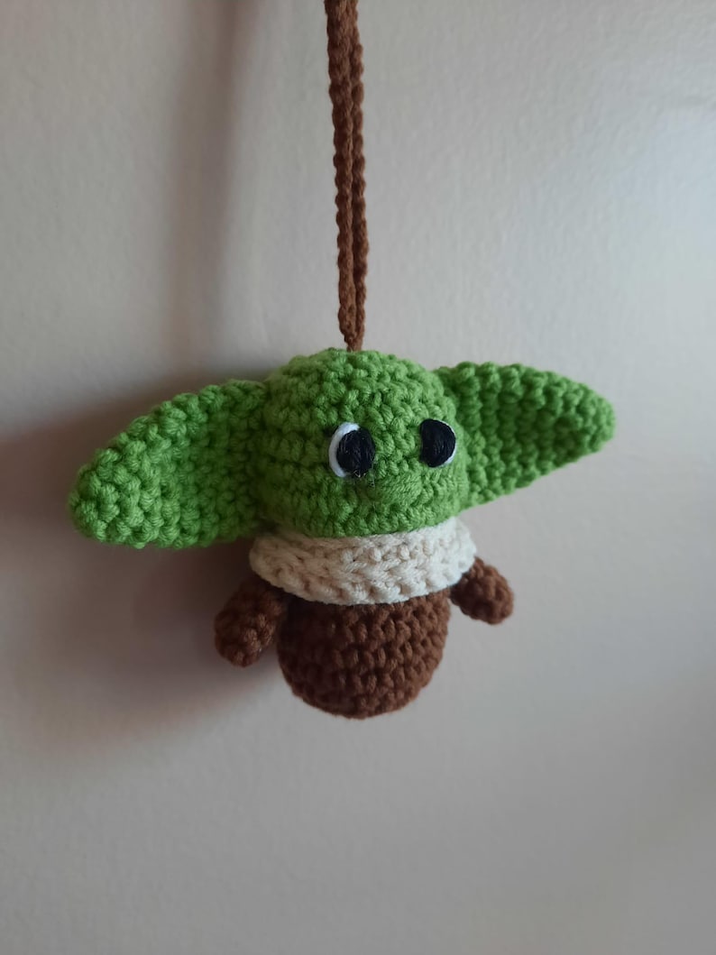May include: A green crocheted Yoda ornament with brown and white details hanging from a brown string.