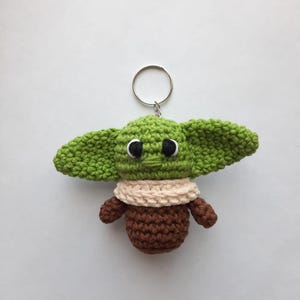May include: Green and brown crocheted keychain shaped like a character from the Star Wars franchise. The keychain has a silver ring.