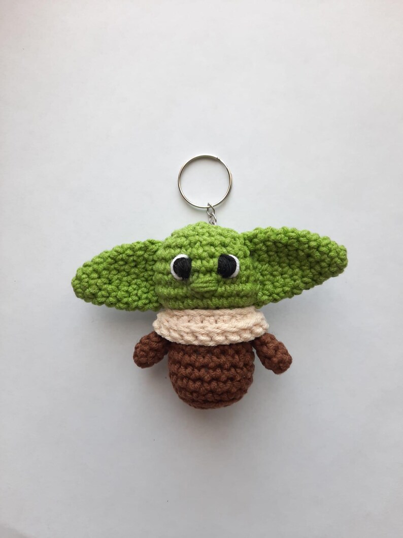 May include: Green and brown crocheted keychain shaped like a character from the Star Wars franchise. The keychain has a silver ring.
