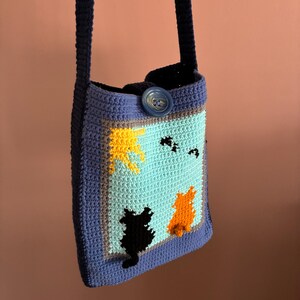 May include: Crocheted shoulder bag with a blue strap and button closure. The bag features a light blue panel with a scene of two cats, a yellow sun, and black birds. The bag is framed with a blue border.