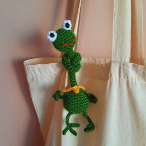 Könnte beinhalten: Eine handgefertigte grüne Häkelfrosch mit weißen und schwarzen Augen, einem roten Mund und einem gelben Kragen. Der Frosch ist an einer beigefarbenen Tragetasche befestigt. Die Arme und Beine des Frosches sind ausgestreckt, was ihm ein verspieltes Aussehen verleiht.