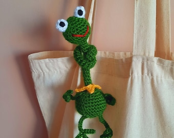 May include: A handmade green crochet frog with white and black eyes, a red mouth, and a yellow collar. The frog is attached to a beige tote bag. The frog's arms and legs are outstretched, giving it a playful appearance.