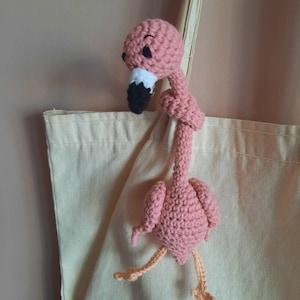 Puede incluir: Un flamenco rosa hecho a mano con detalles de pico negro y blanco y patas color melocotón. El flamenco está sujeto a una bolsa de mano beige. La cabeza del flamenco está girada hacia un lado.