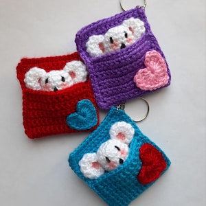 May include: Three crocheted keyrings featuring a white mouse peeking out of a pocket with a heart on the side. The keyrings are in red, purple, and blue.