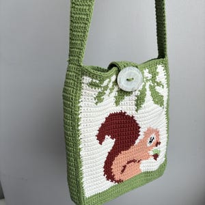 May include: A handmade green crochet shoulder bag with a detailed squirrel design. The bag features a white and green forest pattern, a brown squirrel holding a nut, and a large button closure. The strap and trim are green.