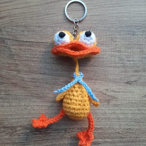Könnte beinhalten: Ein handgefertigter Enten-Schlüsselanhänger aus Häkelarbeit. Die Ente hat einen gelben Körper, einen orangefarbenen Schnabel und Füße sowie große weiße Augen mit schwarzen Pupillen. Ein blauer Schal ist um den Hals gebunden. Der Schlüsselanhänger ist an einem silbernen Metallring befestigt.
