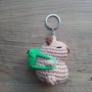May include: A handmade crochet keychain featuring a small, light brown animal with a green accessory. The keychain has a silver ring and a brown heart-shaped detail on top. The animal has black eyes and is made of soft yarn.