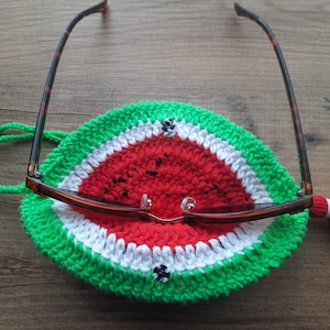 May include: A crocheted eyeglass case in the shape of a watermelon slice. The case features a green rind, a white stripe, and a red center with black seed details. Brown eyeglasses rest on the case.