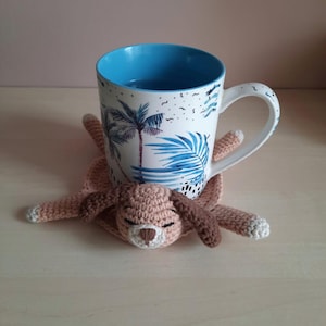 Könnte beinhalten: Eine weiße Keramiktasse mit blauem Innenraum und Palmenmuster steht auf einem gehäkelten, hundeförmigen Untersetzer. Der Untersetzer ist hellbraun mit dunkelbraunen Ohren und Pfoten sowie weißen Akzenten auf den Pfoten.
