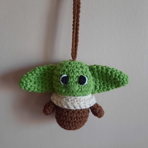 May include: A green crocheted plush toy of a popular character from the Star Wars franchise, with brown crocheted arms and legs, and a white crocheted scarf. The toy is hanging from a brown crocheted string.