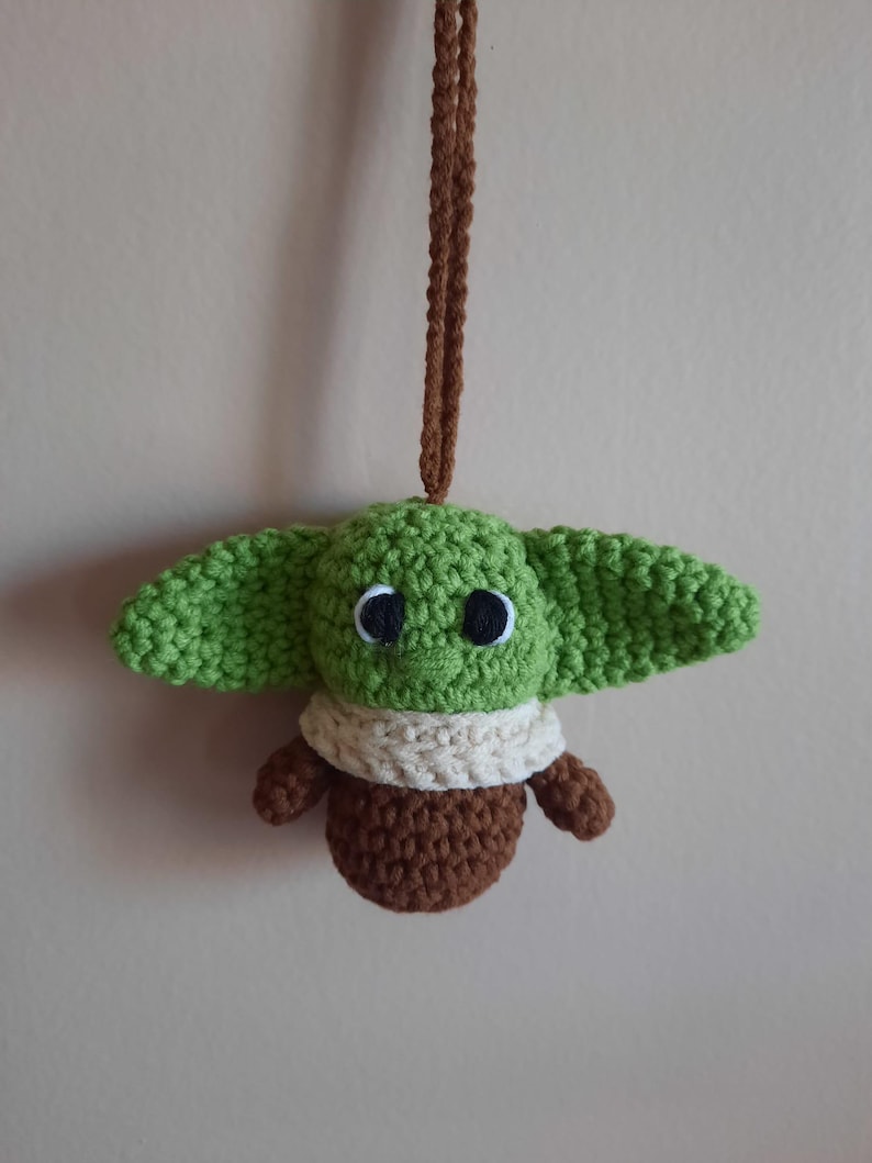 May include: A green crocheted plush toy of a popular character from the Star Wars franchise, with brown crocheted arms and legs, and a white crocheted scarf. The toy is hanging from a brown crocheted string.