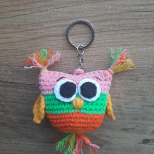 May include: A handmade crochet owl keychain. The owl features a pink head, large white eyes, and a green and orange body. The keychain has a silver metal ring and chain, and colorful yarn accents.