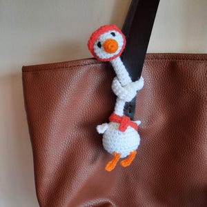 Könnte beinhalten: Eine handgefertigte, weiße Häkelgans mit orangefarbenem Schnabel und Füßen, die einen roten Schal und Hut trägt. Die Gans ist an einem braunen Lederriemen befestigt. Die Tasche hat eine satte, warme braune Farbe.