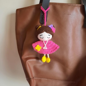 May include: A brown leather tote bag with a crocheted doll keychain. The doll is wearing a pink dress with yellow shoes and a flower.