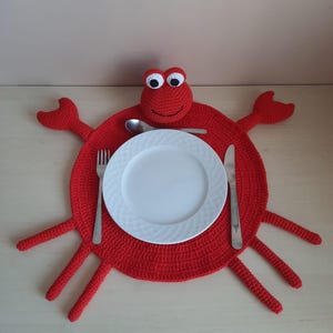 Op de afbeelding: Een rode gehaakte krabvormige placemat met een wit bord, vork, mes en lepel. De krab heeft wiebelogen en uitgestrekte klauwen. De placemat is een grillige toevoeging aan elke eettafel.