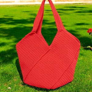 Puede incluir: Una bolsa de mano de crochet de color rojo brillante con una parte superior ancha y abierta y dos asas largas. La bolsa está sentada en un césped verde.
