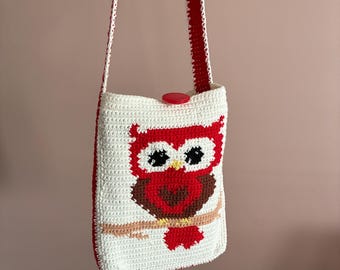 May include: A handmade white crochet shoulder bag featuring a red owl design. The owl has large black eyes and is perched on a brown branch. The bag has a red strap and a red button closure. The bag is approximately 10 inches tall.
