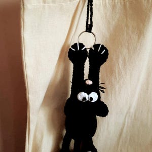 Op de afbeelding: Een zwarte gehaakte katten sleutelhanger met witte accenten op de poten en ogen, ondersteboven hangend. De kat heeft grote, wijde ogen en een kleine roze neus. De sleutelhanger is bevestigd aan een zilveren ring en een zwart koord.