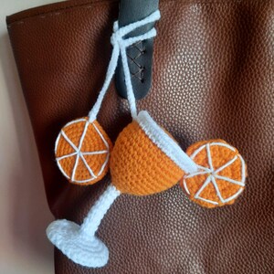 May include: A crocheted cocktail charm in orange, featuring two orange slices. The cocktail glass is orange with a white rim and stem. The charm is suspended from a brown leather bag.