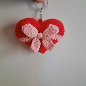May include: A handmade red crochet heart keychain with a pink bow. The heart has a pink border and is attached to a silver keyring. The keychain is a charming accessory.