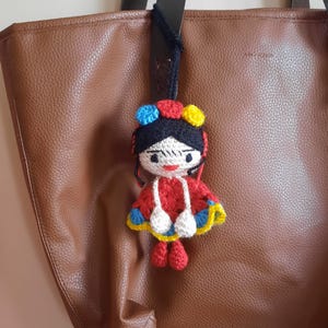 May include: A handmade crochet doll with a red dress, white accents, and a colorful headpiece. The doll is hanging from a brown leather tote bag. The doll's features include a black hair, white face, and red lips.
