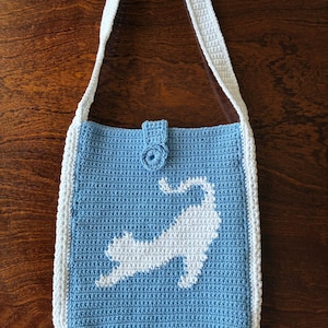 May include: A handmade crochet shoulder bag in light blue with a white cat silhouette. The bag features a white border, strap, and button closure. The bag is approximately 12 inches wide and 14 inches tall.