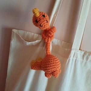 May include: A bright orange crocheted dinosaur toy with yellow detailing, suspended from a cream canvas tote bag. The dinosaur features a smiling face and is crafted from textured yarn. The bag is a natural colour.