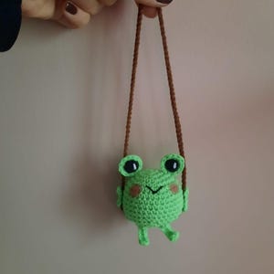 Puede incluir: Una pequeña rana verde tejida a ganchillo, con grandes ojos negros y mejillas rosadas, suspendida por una cuerda marrón. La rana tiene pequeñas patas y brazos, y una boca sonriente. Es un artículo hecho a mano.