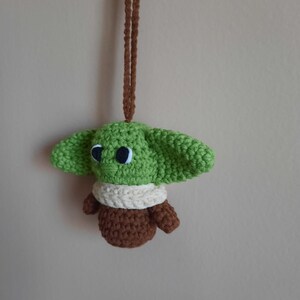 May include: A green crocheted plush toy with brown and white accents hanging from a brown cord. The toy has large eyes and resembles a popular fictional character.