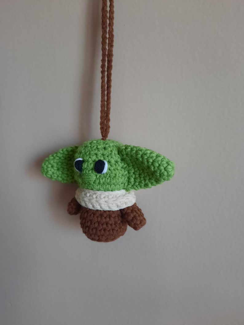 May include: A green crocheted plush toy with brown and white accents hanging from a brown cord. The toy has large eyes and resembles a popular fictional character.