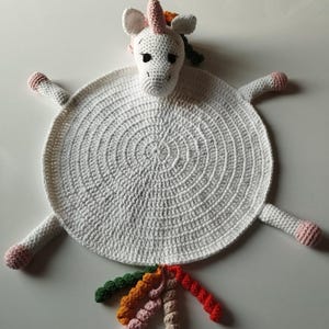 Könnte beinhalten: Weiße gehäkelte Babydecke mit einem Einhorn und einem bunten Regenbogen-Schwanz. Das Einhorn hat rosa Ohren und ein rosa Horn.