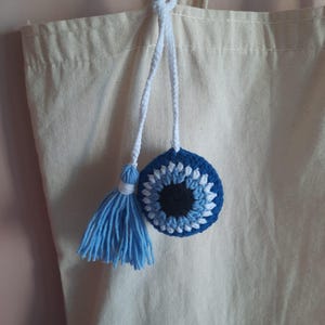 May include: A blue and white crocheted evil eye charm with a matching tassel, hanging from a white cord. The charm is attached to a beige canvas tote bag. The evil eye design features a black center, surrounded by blue and white yarn.