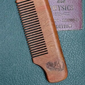 May include: A brown wooden comb with a dark brown handle and a logo on the bottom. The comb has fine teeth and is made of natural wood.