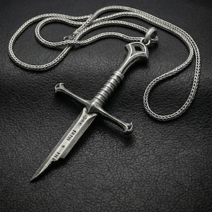 May include: A silver-colored pendant necklace featuring a detailed dagger design. The dagger has a cross-shaped hilt and a blade with etched markings. The necklace chain is a woven style, and the pendant is displayed against a dark background.
