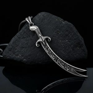May include: A silver-colored pendant necklace featuring a curved sword design. The sword has intricate detailing on the handle and blade, with engraved text. The pendant hangs on a thin chain against a dark, textured background.