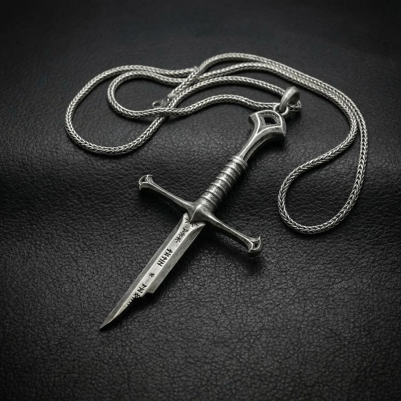 May include: A silver-colored pendant necklace featuring a detailed dagger design. The dagger has a cross-shaped hilt and a blade with etched markings. The necklace chain is a woven style, and the pendant is displayed against a dark background.