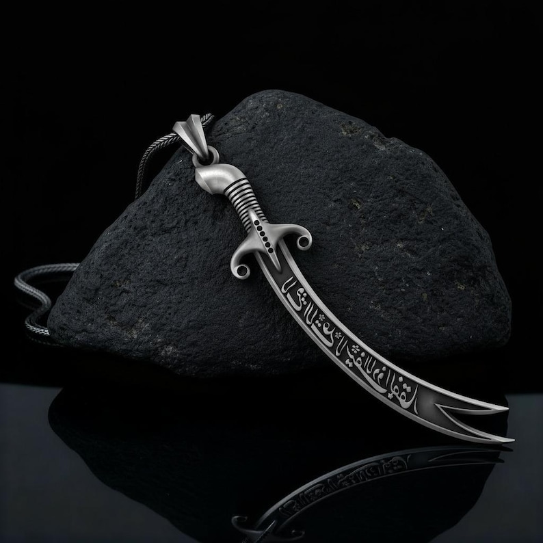 May include: A silver-colored pendant necklace featuring a curved sword design. The sword has intricate detailing on the handle and blade, with engraved text. The pendant hangs on a thin chain against a dark, textured background.