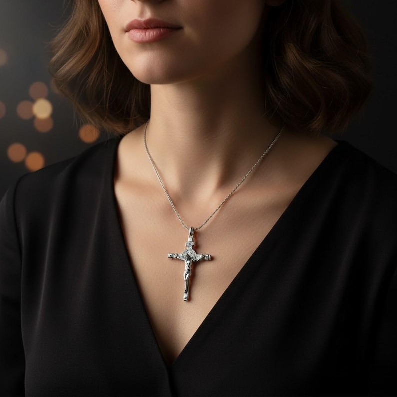 Puede incluir: Un collar con colgante de cruz de plata. El colgante presenta una detallada representaci&oacute;n de Jes&uacute;s en la cruz, con detalles brillantes. El collar se lleva sobre una blusa negra, creando un contraste llamativo. La cadena es delicada y parece ser de plata.
