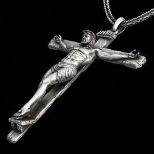 May include: A silver-toned pendant necklace featuring a detailed depiction of Jesus on a cross. The figure is sculpted with intricate detail, and the cross bears the inscription "INRI". The necklace hangs from a silver chain against a black background.