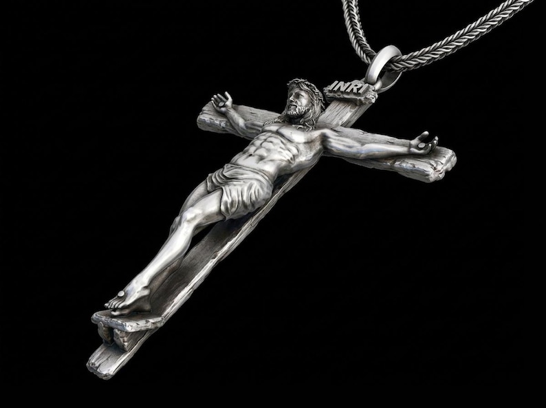 May include: A silver-toned pendant necklace featuring a detailed depiction of Jesus on a cross. The figure is sculpted with intricate detail, and the cross bears the inscription "INRI". The necklace hangs from a silver chain against a black background.