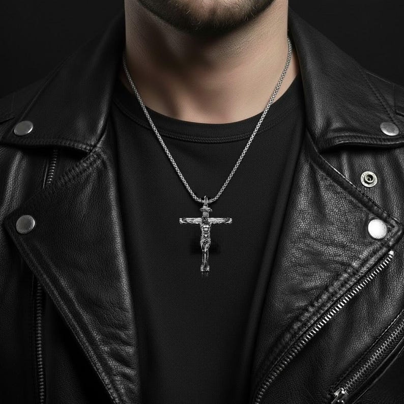 May include: A silver-colored crucifix pendant necklace. The pendant depicts a detailed figure of Jesus Christ on the cross. The necklace is worn over a black t-shirt and a black leather jacket.